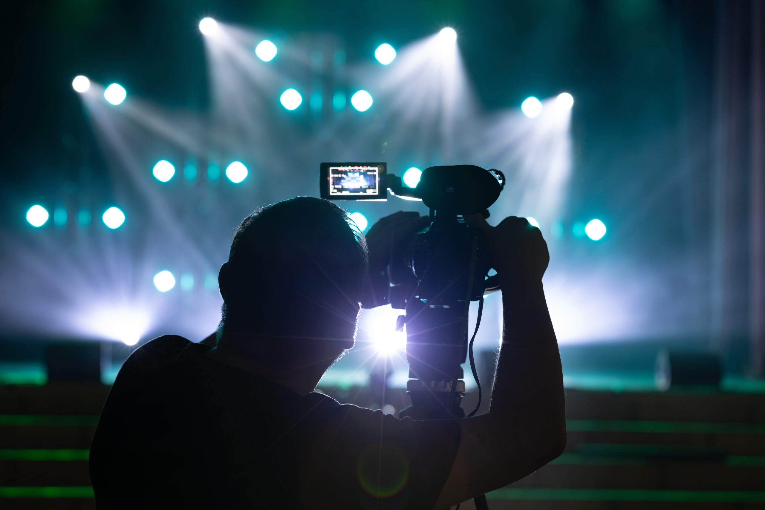 About silhouette of a cameraman operating a video camera on a tripod, capturing a live event under bright blue stage lighting with lens flare and glowing bokeh in the background.